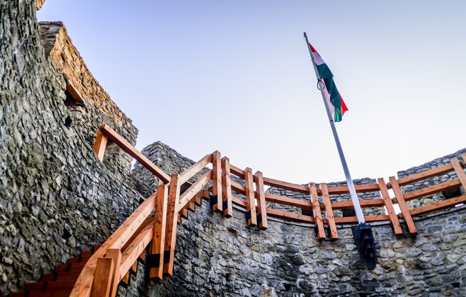 Minden megváltozott Low-angle view of stone castle walls, wooden staircase, and national flag under clear daylight.